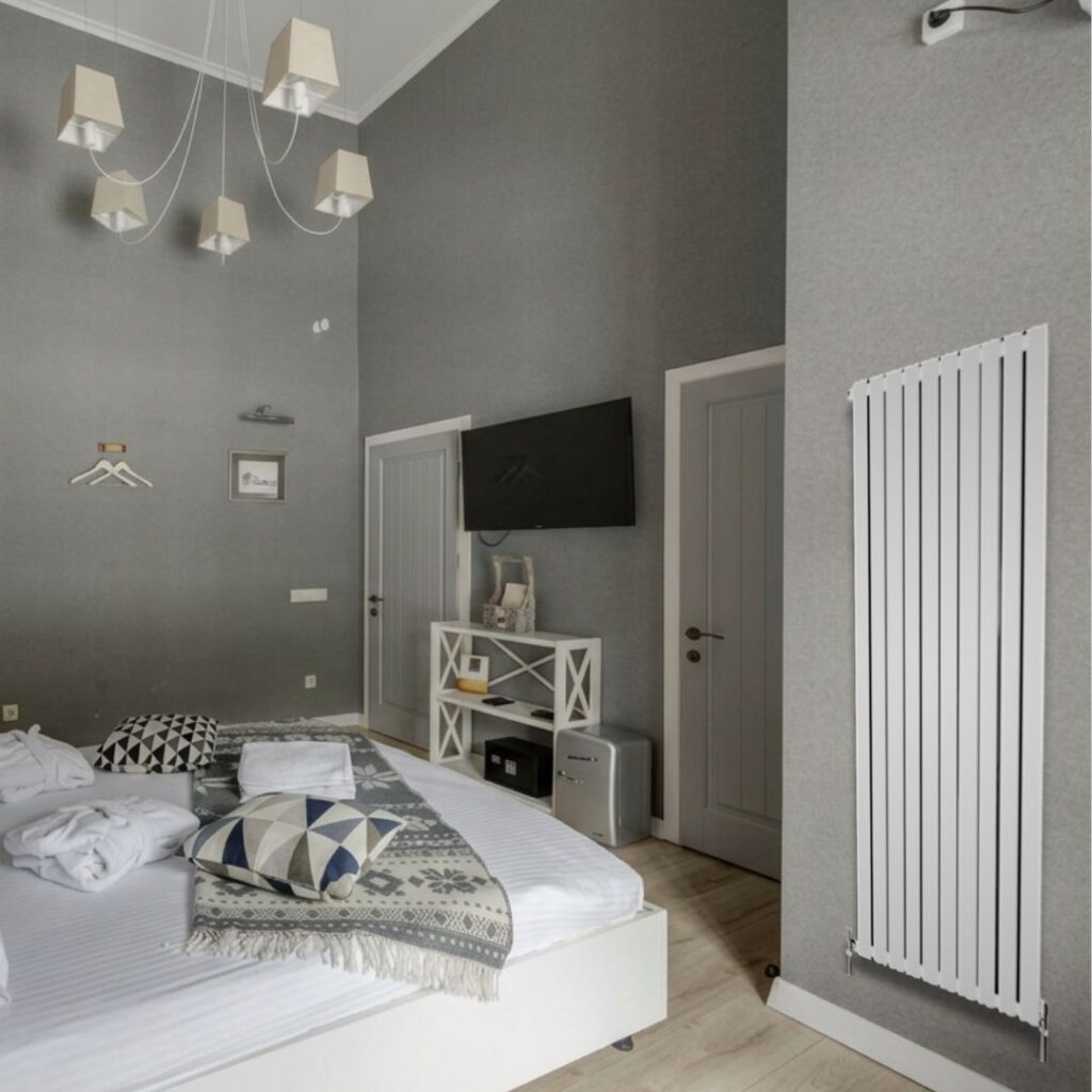 Modern bedroom with a white vertical radiator mounted on a grey wall beside a wall-mounted TV and minimalist décor.